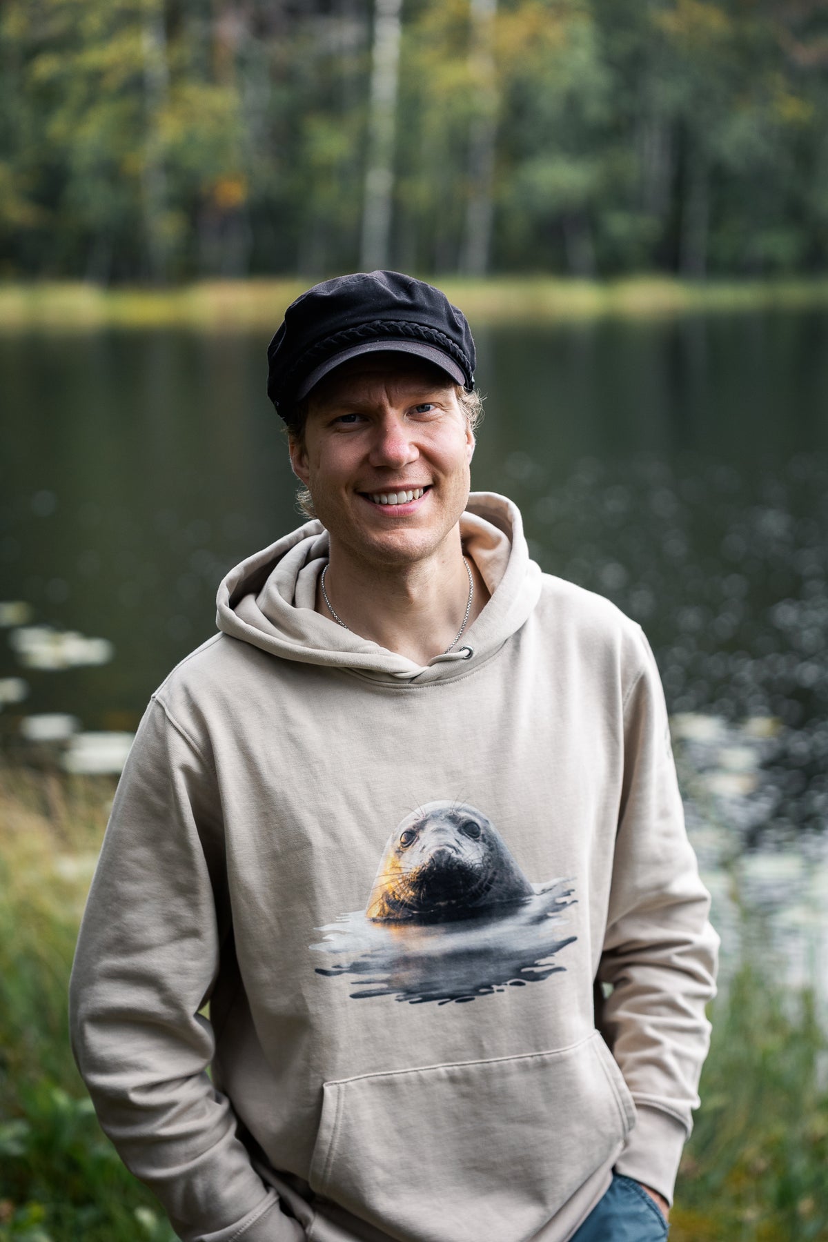 Jetro Luhtaa smiling by a Nordic lake, wearing the sustainable beige organic cotton hoodie by Novapuro featuring a Saimaa ringed seal graphic.