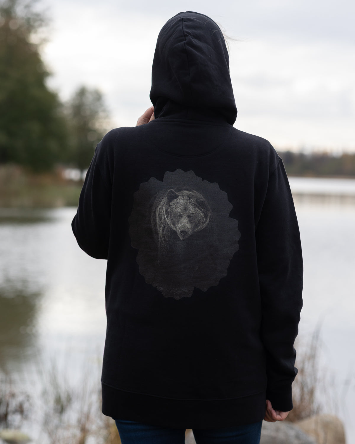 Auli Leivonen wearing a black Novapuro organic cotton hoodie with a majestic Nordic bear photography print on the back, standing outdoors by a lake.