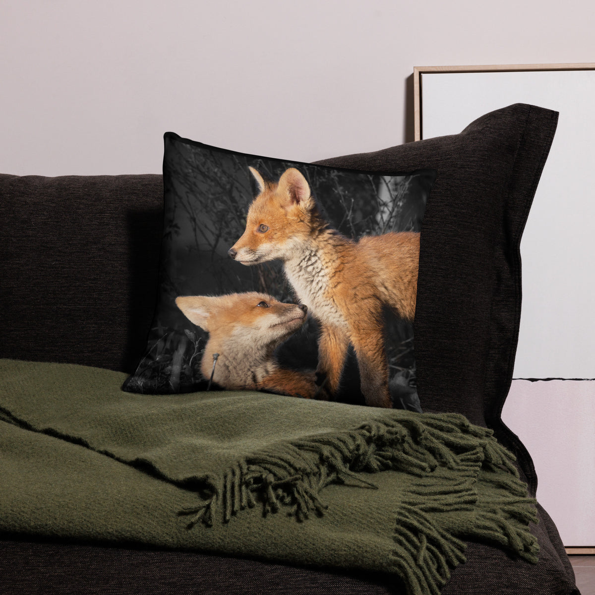 Novapuro x Auli Leivonen dark decorative throw pillow featuring two playful Nordic fox kits, resting on a dark sofa with a green cozy blanket.