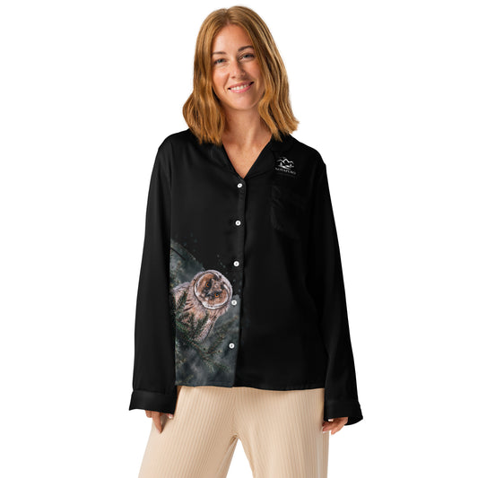 A woman wearing the black Novapuro x Jarno Virtanen long sleeve pajama top featuring a Nordic long-eared owl chick print on the lower side, standing in a cozy room.