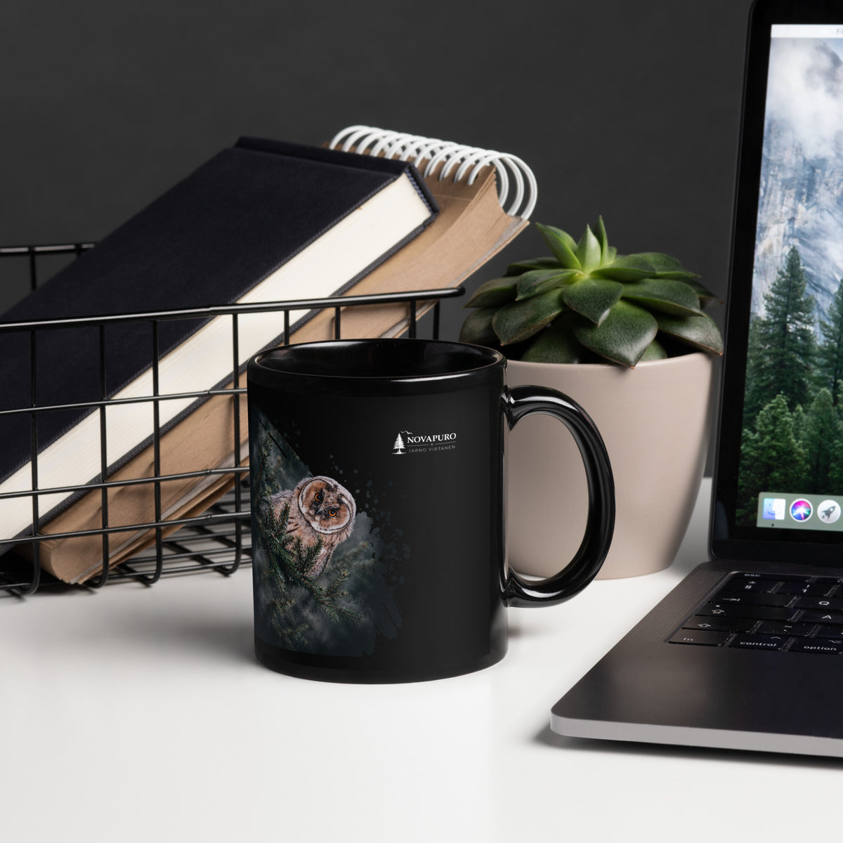 A black glossy ceramic mug featuring a Nordic long-eared owl chick print by Jarno Virtanen, sitting on a desk next to a notebook.