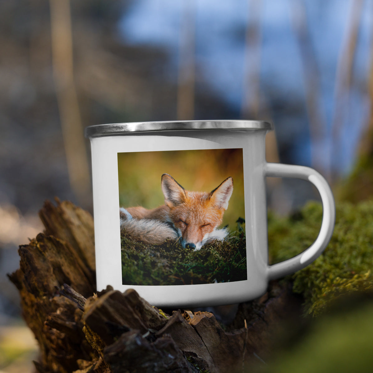 A white enamel camper mug with a silver rim featuring an original photograph of a resting red fox by Mikko Monto, set in a rustic outdoor environment.
