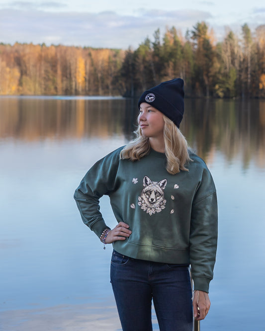 A young woman modeling the green Annette Thomé-Marttinen x Novapuro Nordic Fox organic oversize sweatshirt and a black beanie, standing by a calm Nordic lake in autumn.
