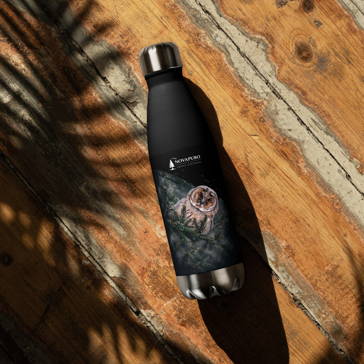 A black double-walled stainless steel water bottle featuring a Nordic long-eared owl chick print by Jarno Virtanen, resting on a wooden surface.