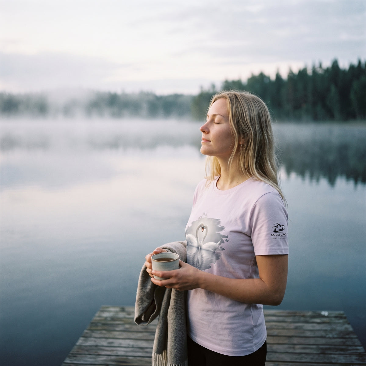A woman standing peacefully on a wooden pier by a misty Nordic lake, wearing a pale pink sustainable women's organic cotton t-shirt featuring a swan wildlife print and sleeve logo.