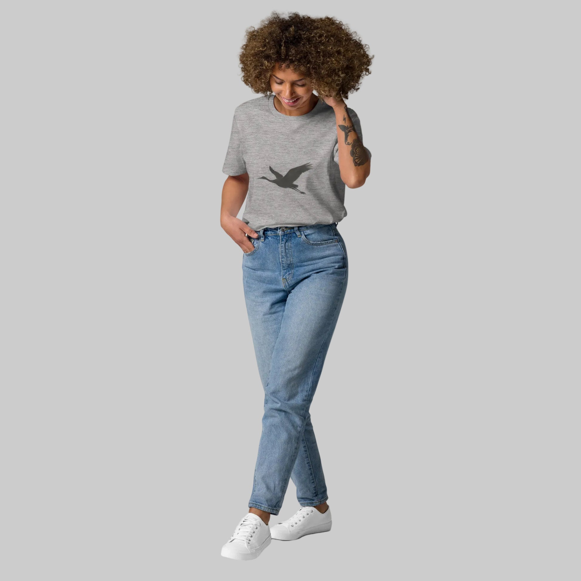 A female model wearing a heather grey Novapuro organic cotton t-shirt with a minimalist black flying bird design, styled with blue jeans.